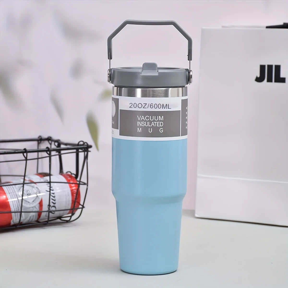 Vacuum-insulated stainless steel tumbler in light blue color, with a handle, placed on a table alongside a wire basket and a white box
